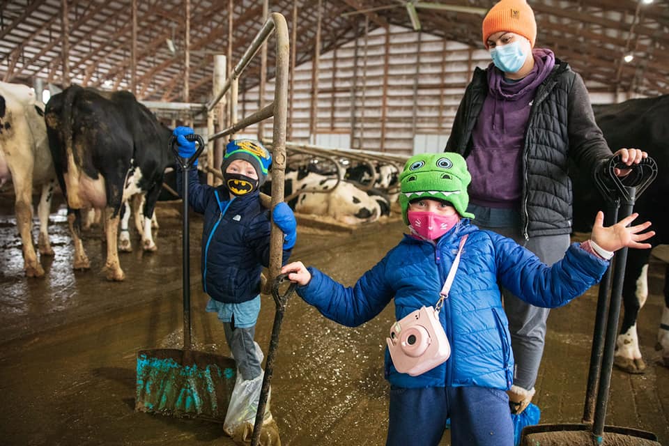 two children and an adult standing in a cow barn with shovels