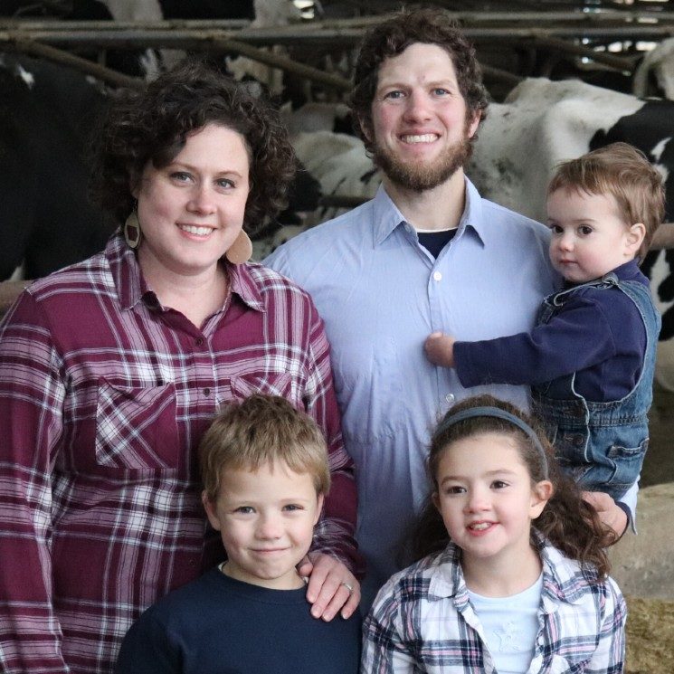 woman in marron plaid, man in blue button shirt holding baby, girl & boy standing in front of them in a dairy barn