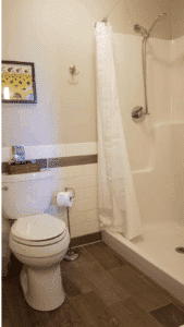 Modern bathroom with white shower and curtain, toilet, beige walls, wood-look tile floor, and yellow framed artwork