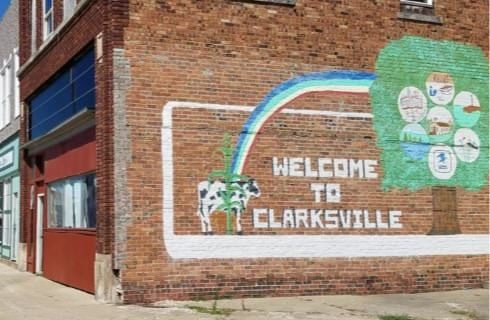 Street level view of main street shops with a large red brick wall decorated with a painting of a black and white cow, rainbow, green tree, and Welcome to Clarksville text