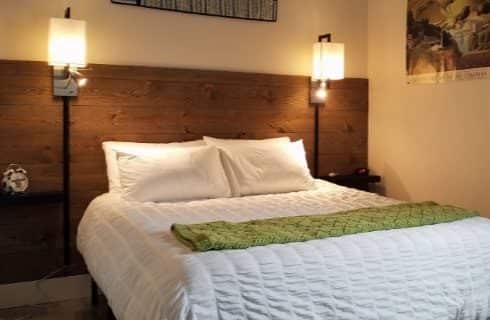 Bed with white bedding and green blanket, dark wooden slats on wall as accent and headboard, wall sconces, and artwork on the walls