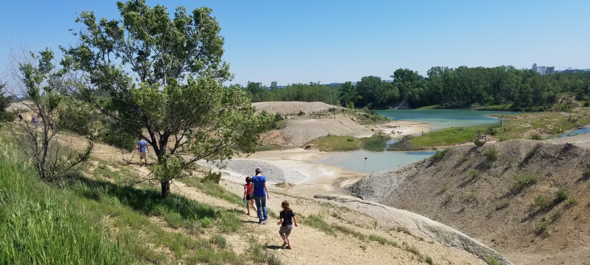 group of people hiking on gravel down to crystal blue water with trees along trail and in basin