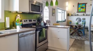 View of a kitchen with white cabinets, green backsplash, and stainless steel appliances and view into dining area with wooden table and green, yellow, and blue chairs