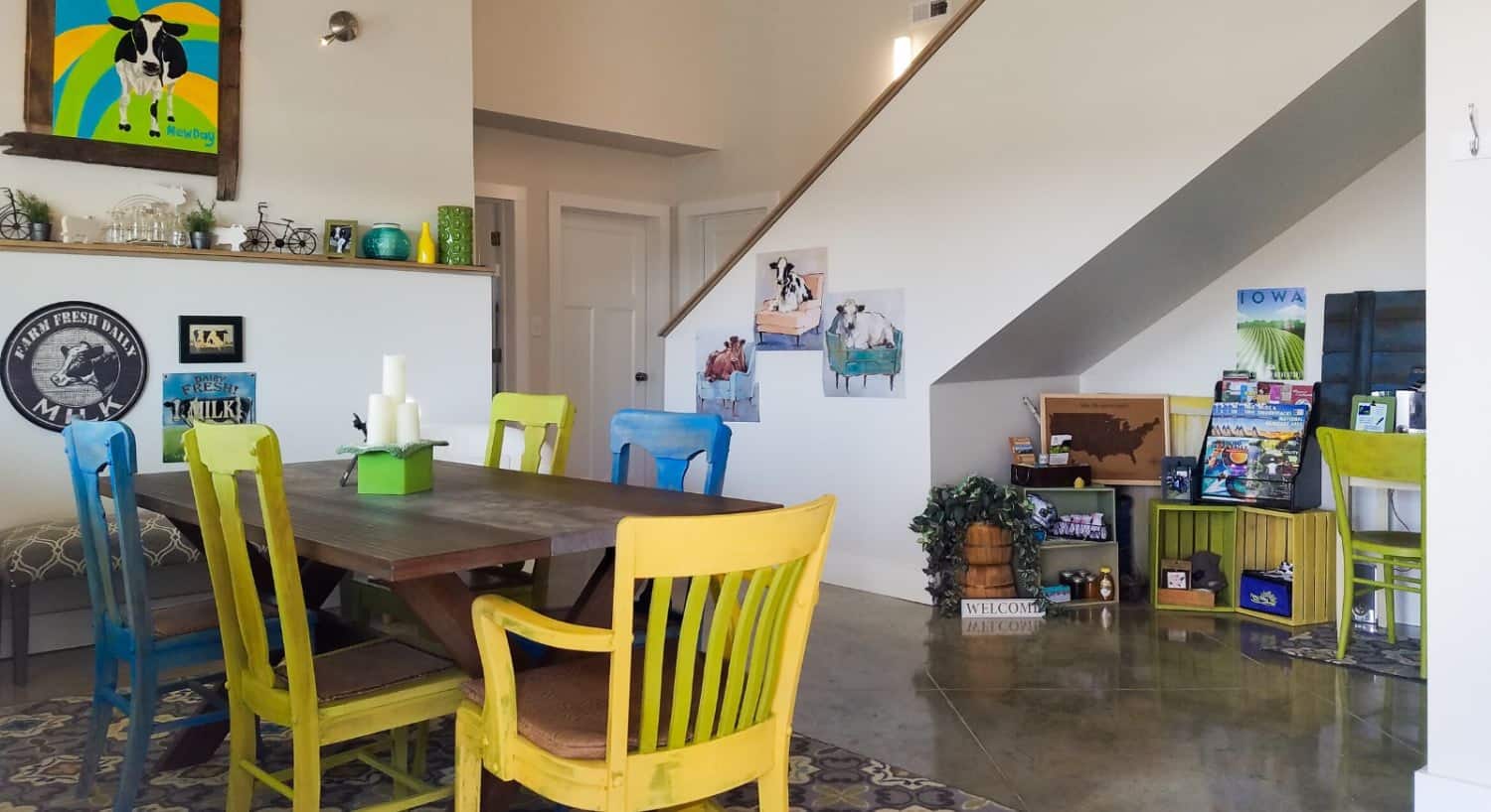 View of dining area with wooden table and blue and yellow chairs, a small nook with old crates storing items for sale, stairs leading upstairs, and cow artwork on the walls