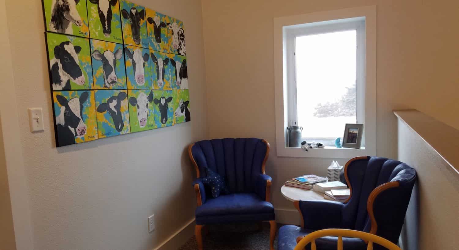 Loft area at the top of the stairs with white walls and trim, carpeting, small sitting area with a table and blue upholstered chairs, and a large cow artwork on the wall