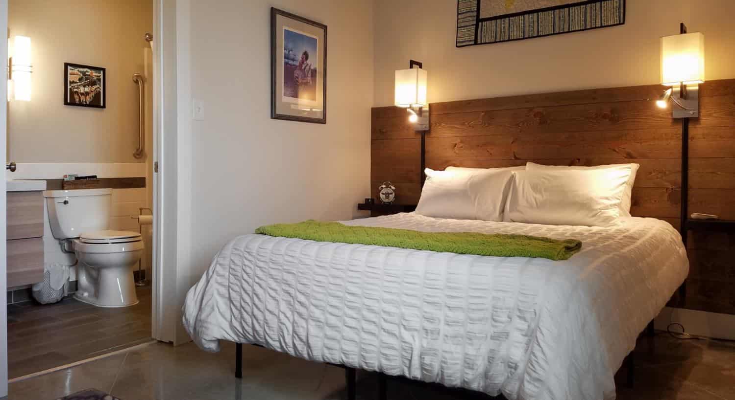 Bed with white bedding and green blanket, dark wooden slats on wall as accent and headboard, wall sconces, artwork on the walls, and view into bathroom