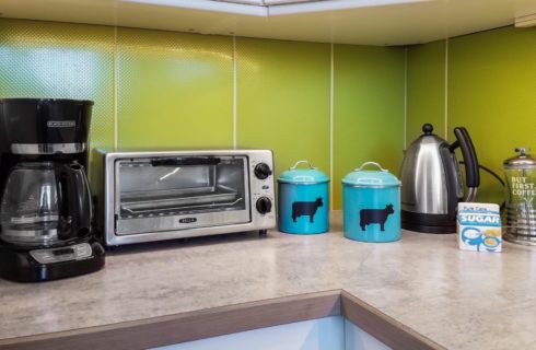 Close up view of a kitchen counter with green backsplash, coffee maker, toaster oven, teapot, and two turquoise containers with black dairy cows