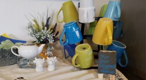 Close up view of a coffee cup stand with blue, green, yellow, and white cups, white cream dish, and white cow head salt and sugar shakers