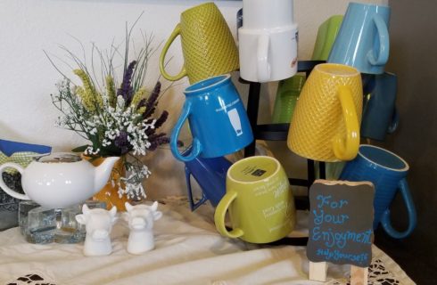 Close up view of a coffee cup stand with blue, green, yellow, and white cups, white cream dish, and white cow head salt and sugar shakers