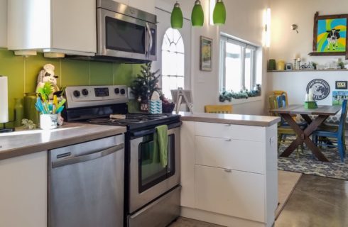 View of a kitchen with white cabinets, green backsplash, and stainless steel appliances and view into dining area with wooden table and green, yellow, and blue chairs