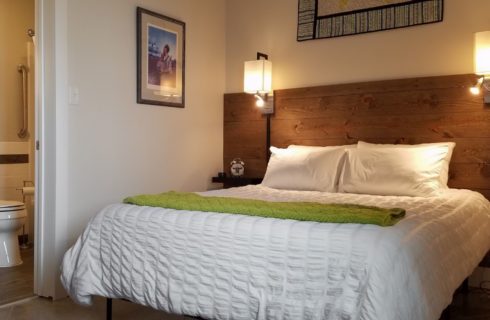 Bed with white bedding and green blanket, dark wooden slats on wall as accent and headboard, wall sconces, artwork on the walls, and view into bathroom