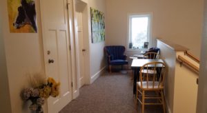 Loft area at the top of the stairs with white walls and trim, carpeting, small table with two chairs and small sitting area with a table and blue upholstered chairs