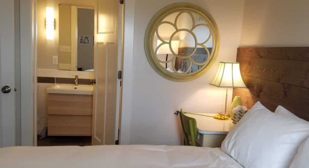 looking into attached bathroom with queen bed and bedside desk with lamp & mirror on wall