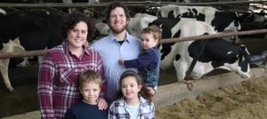 Family with coordinating shirts in solids and plaids in burgundy, navy, light blue, and white hues standing in front of large white and black dairy cows