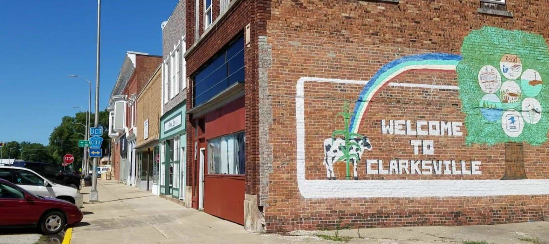 Street level view of main street shops with a large red brick wall decorated with a painting of a black and white cow, rainbow, green tree, and Welcome to Clarksville text