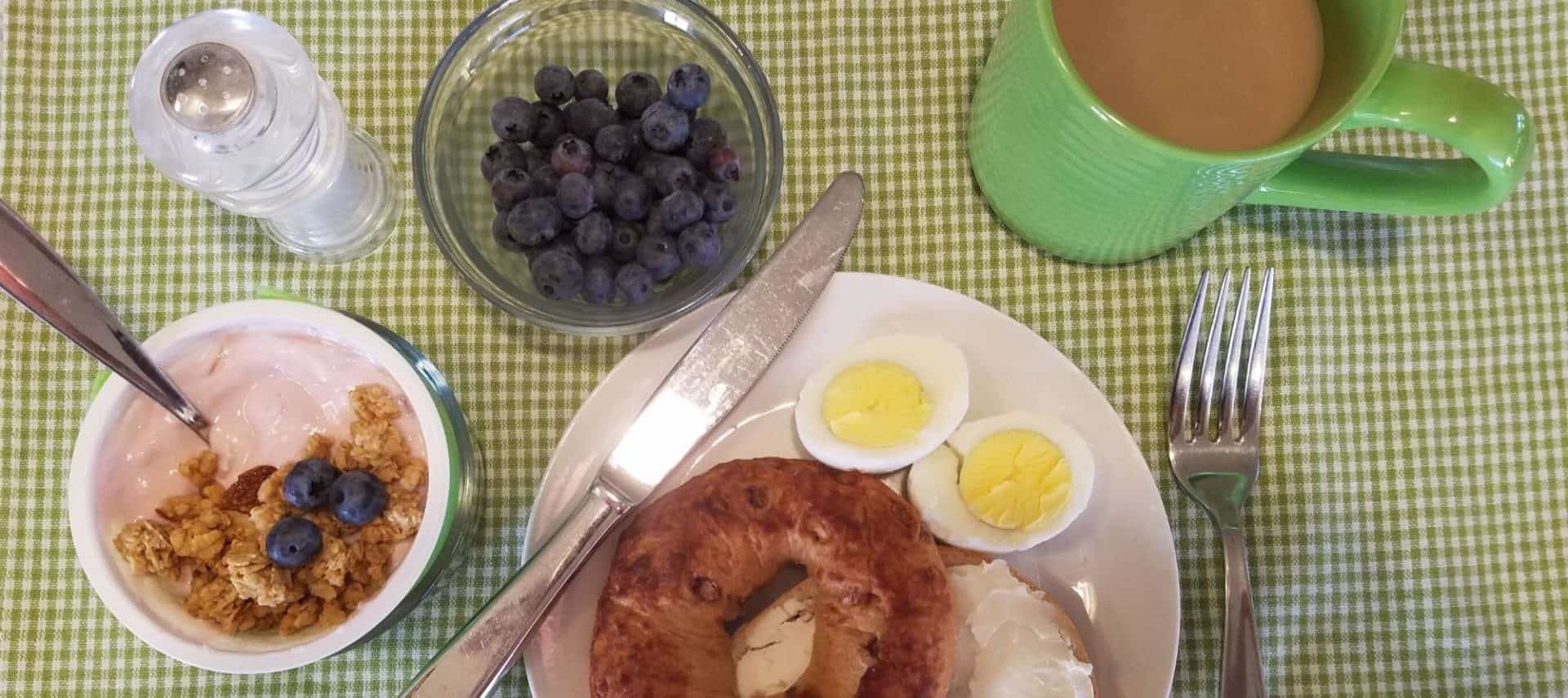 View from above looking down at a bagel with cream cheese, hard boiled egg cut in half, yogurt with granola and blueberries, bowl of blueberries, and green cup with coffee