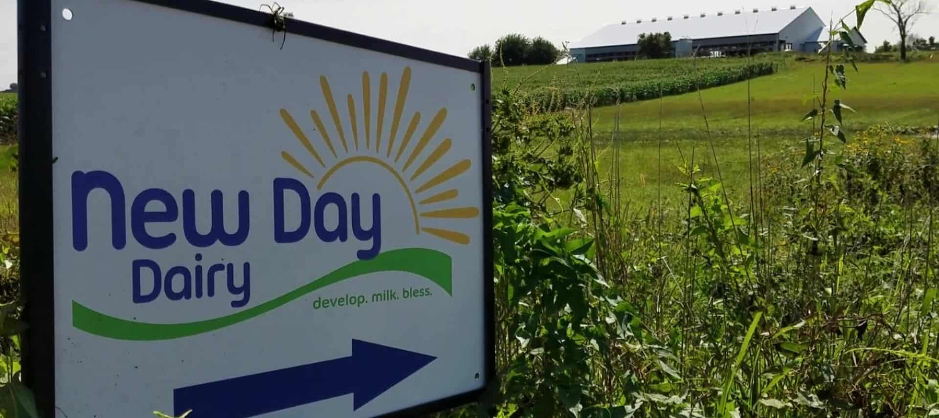 Large sign for New Day Dairy sitting in a large green field with the dairy barn in the background