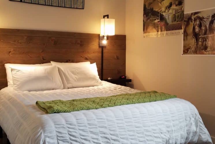 Bed with white bedding and green blanket, dark wooden slats on wall as accent and headboard, wall sconces, and artwork on the walls