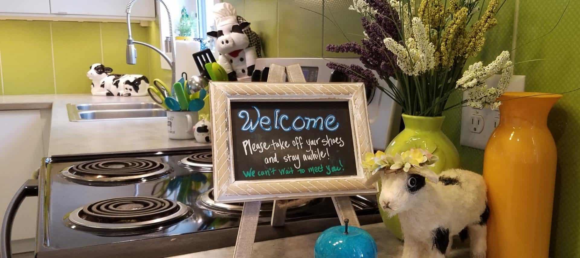 Close up view of countertop in a kitchen with a small Welcome sign, blue glass apple, orange and green vases, and stuffed black and white cow