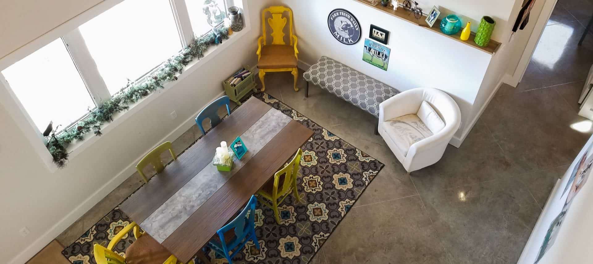 View of a dining area and sitting area from up high with large wooden table, blue and yellow chairs, large coordinating area rug, and upholstered bench and chair