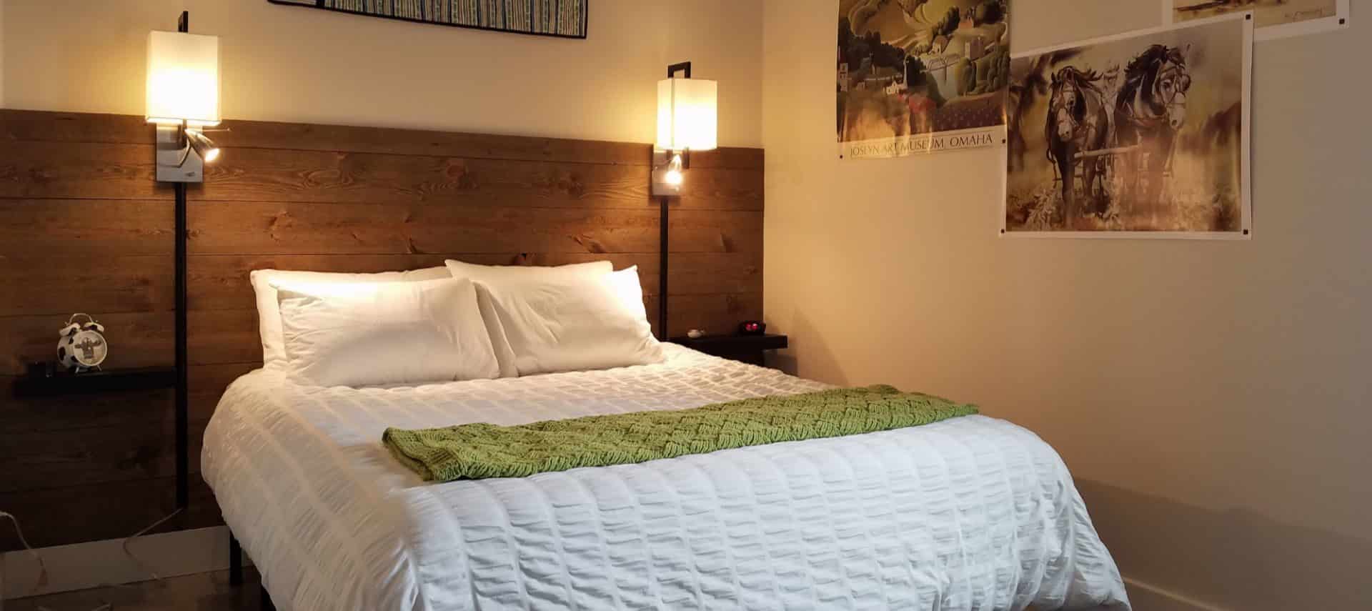 Bed with white bedding and green blanket, dark wooden slats on wall as accent and headboard, wall sconces, and artwork on the walls