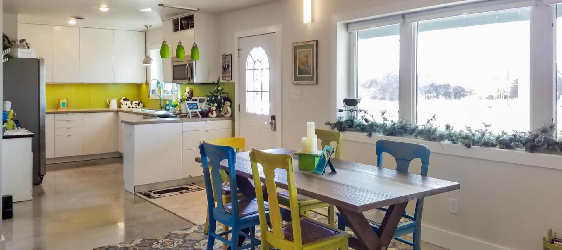 Large dining area and kitchen with wooden table and blue and yellow chairs, view to the outside, white cabinets, and light green backsplash