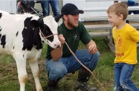 Man with green shirt holding a rope attached to a baby black and white calf for a kid with a yellow shirt