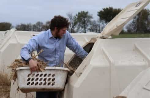 Man with blue shirt holding basket of silage next to large storage containers